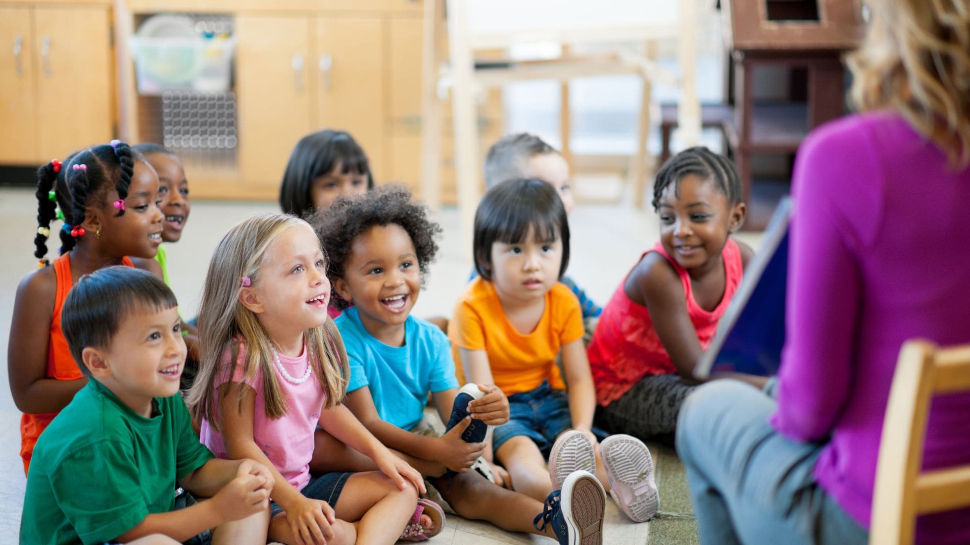 A diverse group of young children sits on the floor in a classroom, attentively listening and smiling at a person out of frame, likely a teacher or storyteller. The children are dressed in colorful clothing and appear engaged and happy, suggesting an interactive learning or storytime session. The background shows classroom cabinets and educational materials, indicating a preschool or early childhood education setting. The atmosphere is warm and inviting, emphasizing a positive, inclusive learning environment for young children.