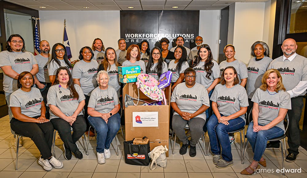 A group of 20 diverse individuals in matching gray shirts with a city skyline design pose in front of a 'Workforce Solutions Greater Dallas' sign, surrounded by donation boxes of diapers and toys. The setting is an office lobby with a Texas flag in the background.