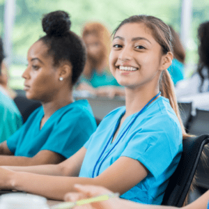 The image shows a group of nursing students in a classroom setting, with one student in the foreground smiling at the camera. They are all wearing teal scrubs, a common uniform for healthcare training programs.