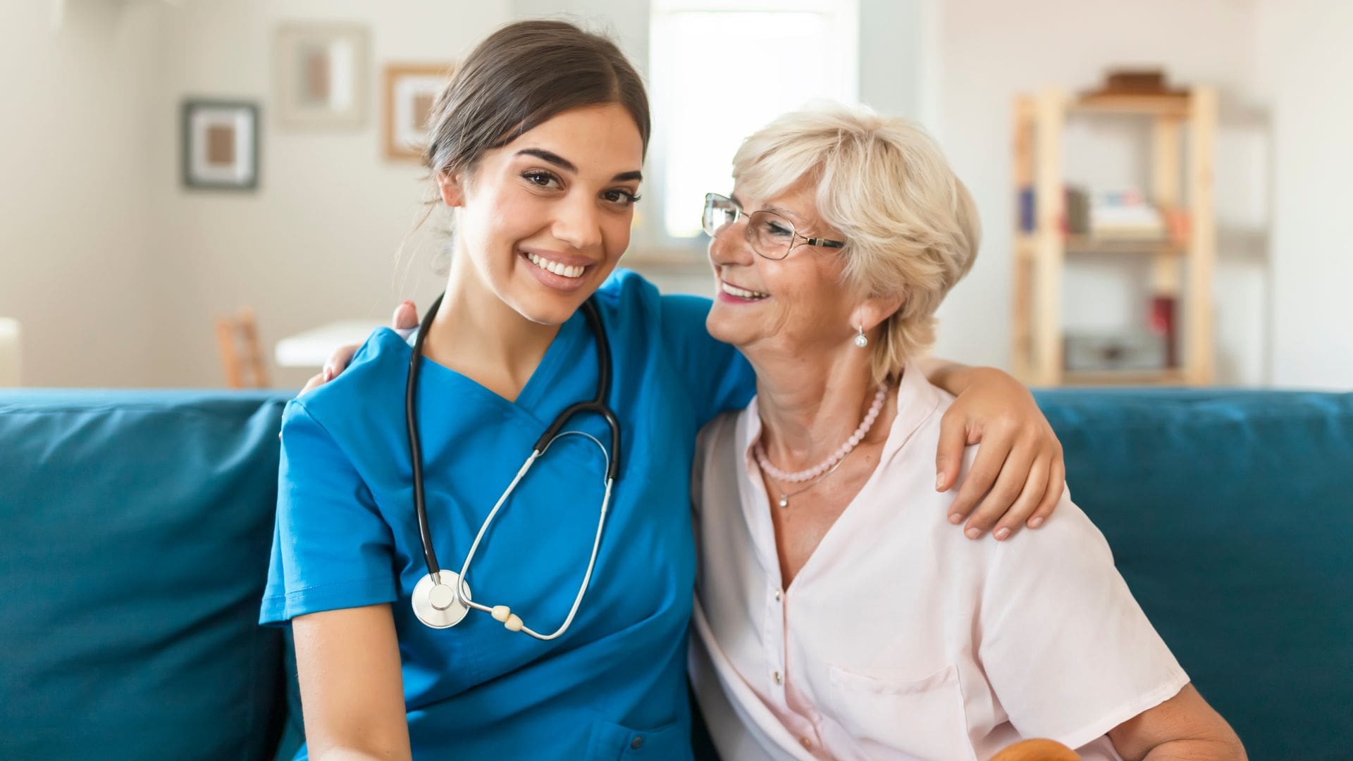 A young female healthcare professional in blue scrubs, with a stethoscope around her neck, sits on a couch and smiles warmly at the camera. She has her arm around an elderly woman with short blonde hair and glasses, who is smiling back at her. The elderly woman is wearing a light pink blouse and pearl earrings, conveying a friendly and caring relationship. The setting appears to be a cozy living room, suggesting a home healthcare or caregiving environment. The image reflects warmth, compassion, and support in healthcare services.