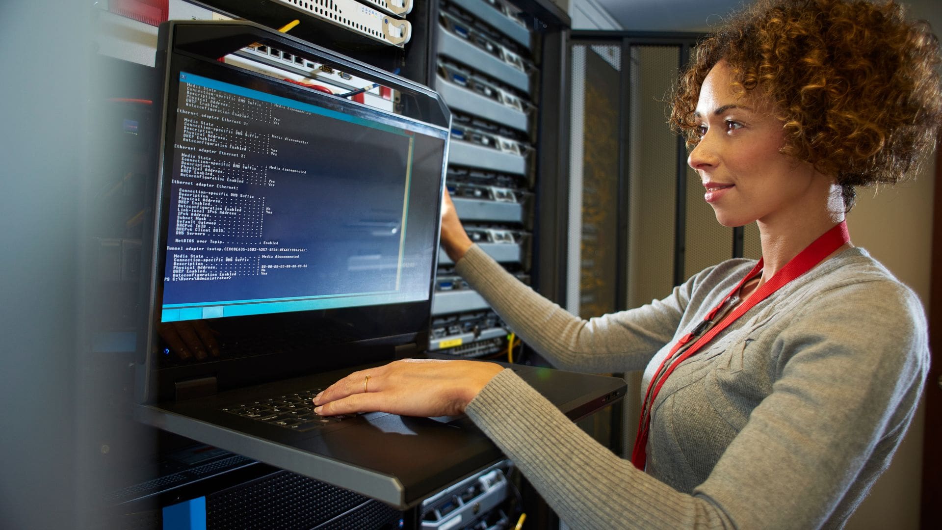 A woman with curly hair wearing a gray sweater and a red lanyard works on a laptop mounted on a rack in a server room. She is focused on the screen, which displays network configuration and diagnostic information. Surrounding her are rows of server racks filled with equipment and cables, indicating a technical and data-centric environment. The image conveys a sense of professionalism and technical expertise in information technology or network management.