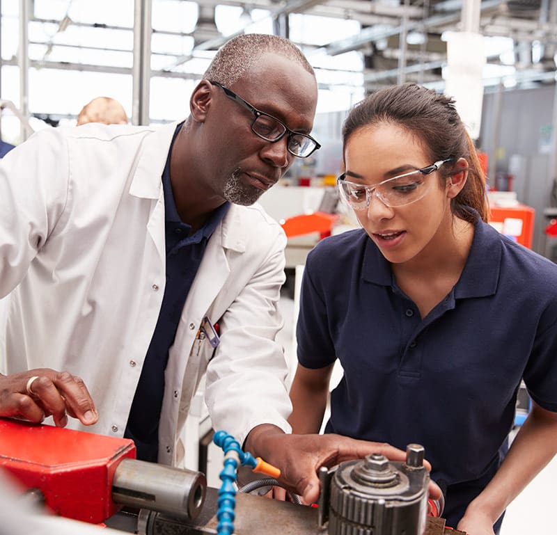 An older man wearing glasses and a lab coat is guiding a younger woman wearing safety goggles and a navy polo shirt in a workshop or industrial setting. They are both focused on a piece of machinery, with the man pointing at a component while explaining its function. The woman is listening attentively, indicating an apprenticeship or hands-on training session. The background shows various tools and equipment, emphasizing a learning environment in a technical or manufacturing field.