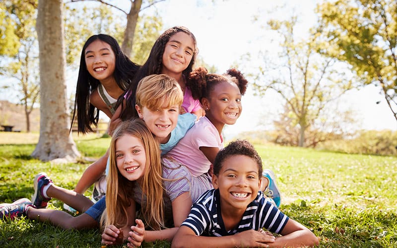 A diverse group of six children is playfully piled on top of each other on the grass in a park. They are all smiling and laughing, showing joy and camaraderie. The children, who are of various ethnic backgrounds, are dressed in casual summer clothes, and the setting is a sunny day with trees and greenery in the background.