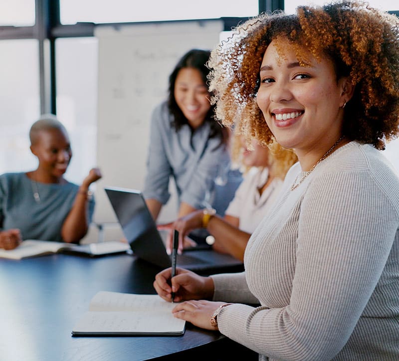 A group of four diverse women are gathered around a table in a bright, modern office setting, engaged in a collaborative discussion. The woman in the foreground, with curly hair and wearing a light sweater, is smiling at the camera while taking notes in a notebook. The other three women in the background are looking at a laptop and a whiteboard, actively participating in the conversation. The atmosphere is friendly and focused, indicating a team meeting, brainstorming session, or employee training.