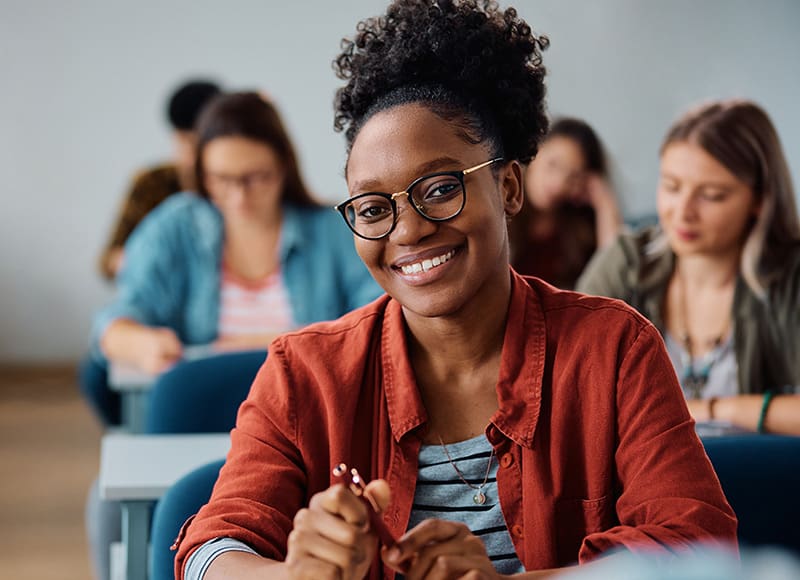 A young woman wearing glasses and a red shirt smiles while sitting at a desk in a classroom, with other students working in the background.