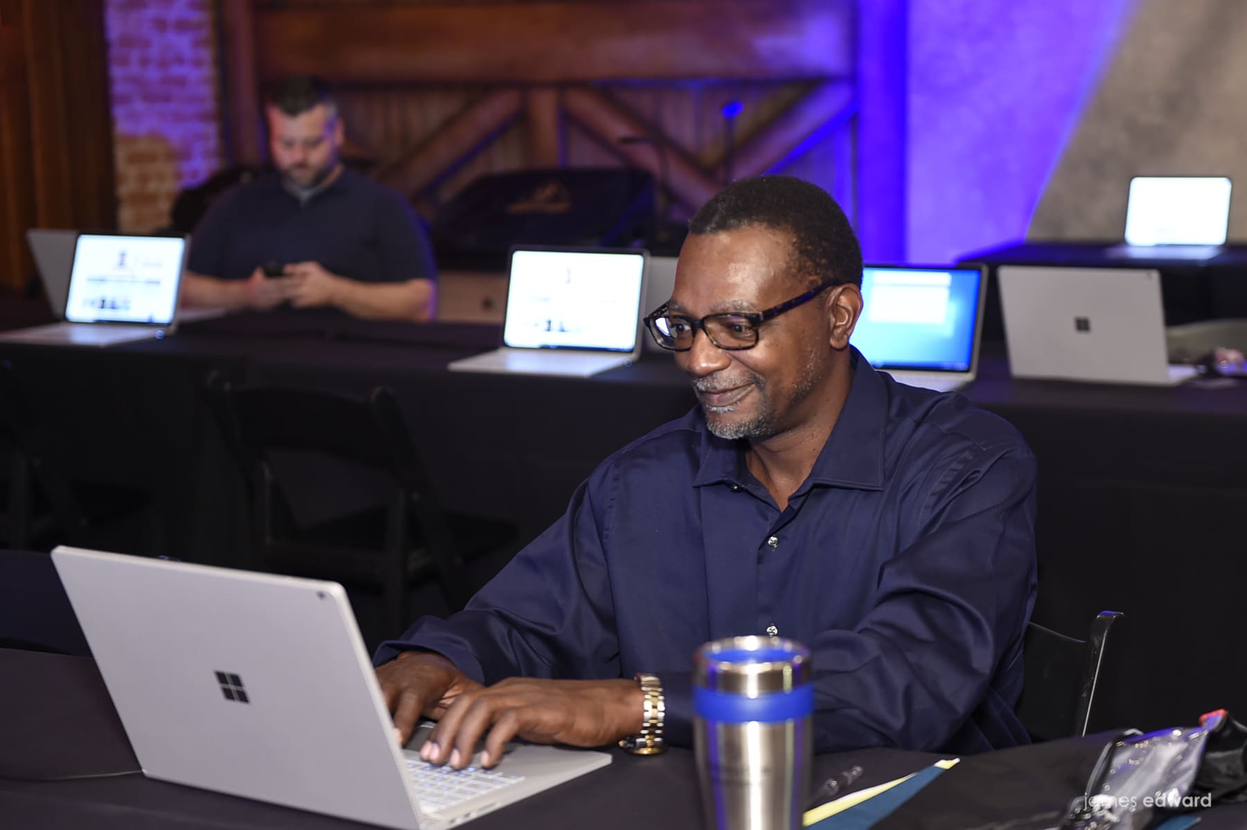 A man wearing glasses and a dark blue shirt is sitting at a table, focused on his laptop, typing. He appears to be engaged in work or a study session. In the background, another man is seated, looking at his smartphone, with several laptops open on the table between them.