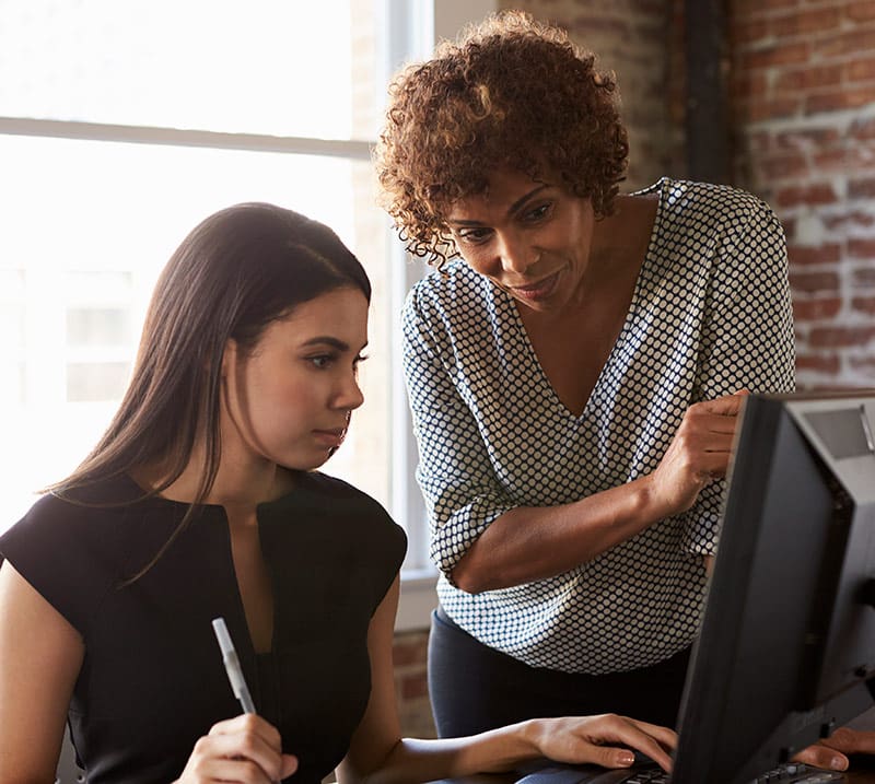 A young woman, likely an intern, is seated at a desk in front of a computer monitor, focused intently on the screen. She is holding a pen and appears engaged in a task or project. Standing beside her is an older woman, possibly a mentor or supervisor, who is leaning in to provide guidance or instructions. The mentor is pointing at the screen, indicating a collaborative effort. Both women are dressed in professional attire, and the setting is an office with a brick wall and large window, creating a warm, well-lit environment.