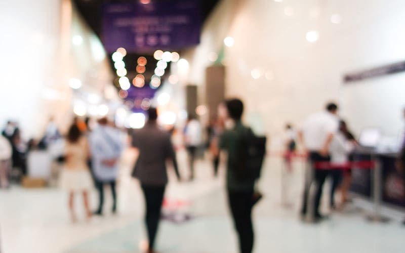 A blurred image of people walking through a busy, well-lit indoor hallway, suggesting a bustling environment such as a job fair or conference.