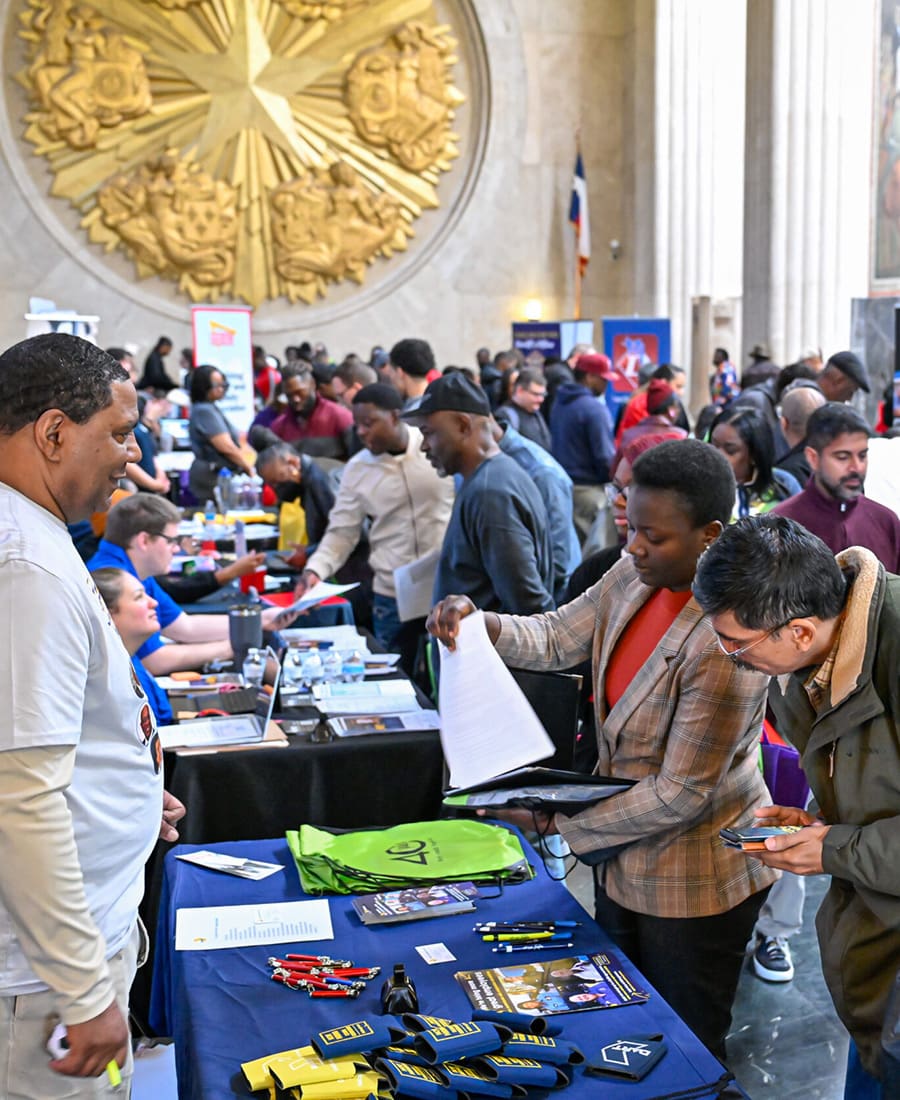 A bustling job fair inside a large hall with a prominent golden star emblem on the wall in the background. Multiple attendees engage with recruiters at various booths lined with promotional materials and informational pamphlets. In the foreground, a man in a white shirt interacts with a woman in a plaid blazer holding documents, while a man beside her checks his phone. The event appears lively, with numerous conversations and exchanges taking place.