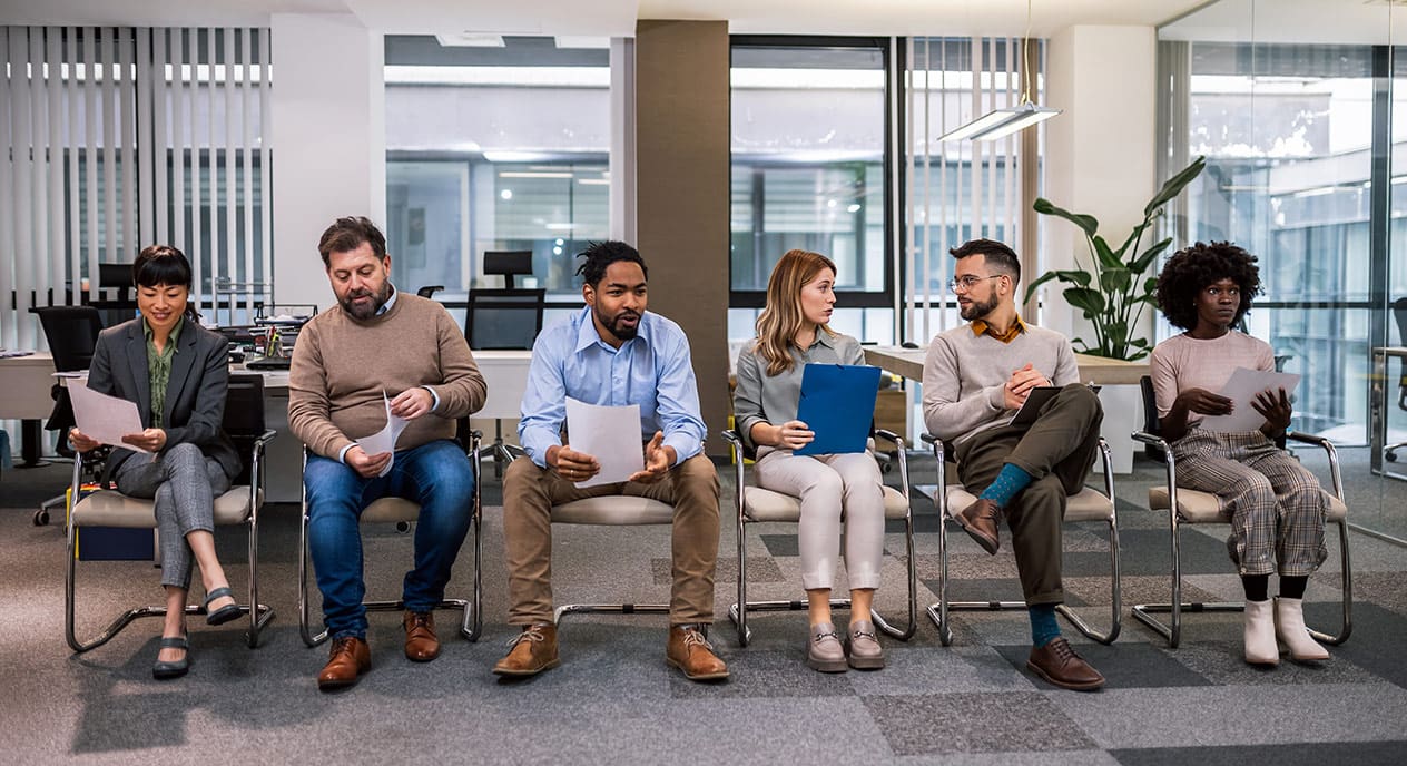 A diverse group of six people are seated in a modern office waiting area, each holding papers or a folder, suggesting they are preparing for a job interview or a meeting. The individuals, who vary in age, gender, and ethnicity, appear focused and contemplative, some reading their documents while others engage in conversation. The setting is professional, with large windows, desks, and office plants in the background, creating a bright and airy atmosphere.