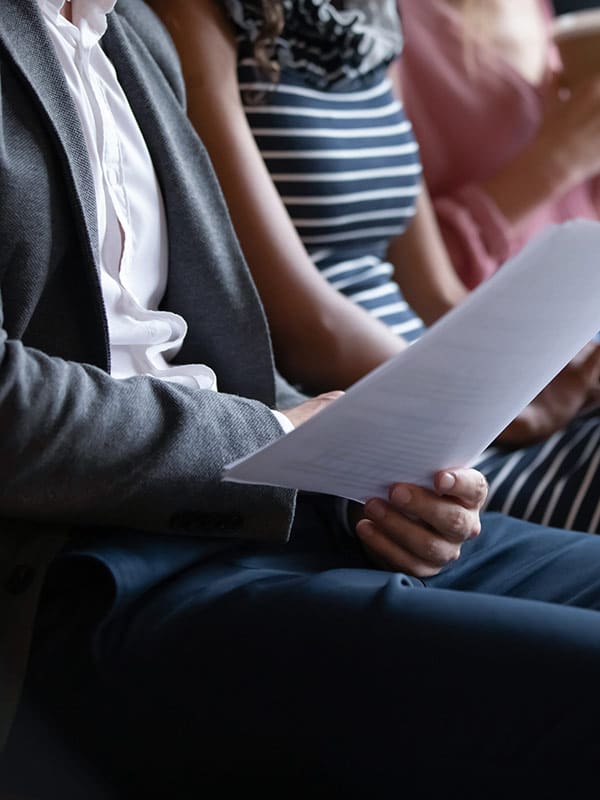 A close-up of a seated individual in a suit holding a document, possibly a resume or an application form, during a professional event or interview session. The person's hand is gripping the paper, showing focus and anticipation. Next to them, a person wearing a striped dress is also visible, suggesting a diverse group of attendees.