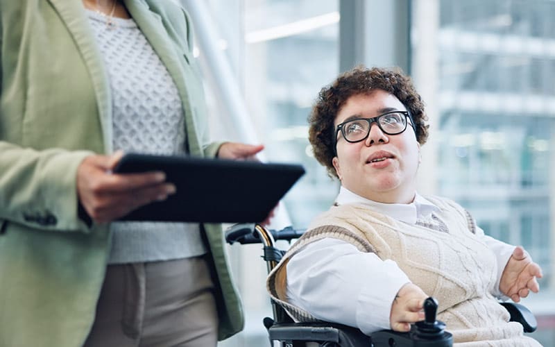 A person with short curly hair and glasses is sitting in a motorized wheelchair, looking attentively towards someone standing beside them. The person standing is holding a tablet and wearing a light-colored blazer over a patterned sweater. They are both in a bright, modern office space with large windows in the background.