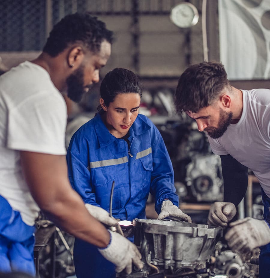 Three mechanics are focused on a task in an auto repair workshop. A woman in the center, wearing a blue jumpsuit and gloves, is intently examining a car part with two men, one on each side. Both men are wearing white t-shirts and gloves, closely inspecting the component and collaborating with the woman. The background features various car parts and tools, indicating a hands-on, technical environment.