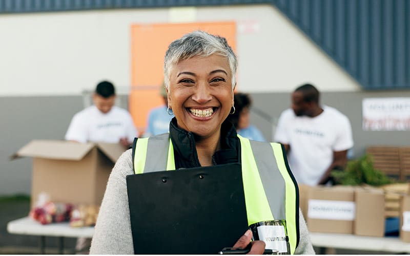 A smiling woman with short gray hair is wearing a high-visibility safety vest and holding a clipboard while standing outdoors. She appears to be at a community event or volunteer activity, with other volunteers working at a table in the background, handling boxes and bags of goods.