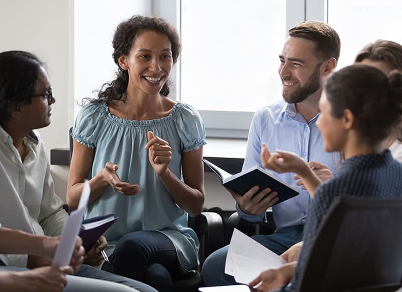 A diverse group of individuals sitting in a circle engaged in a lively discussion. A woman in a light blue blouse is smiling and speaking, holding a notepad, while others listen attentively and hold notebooks.