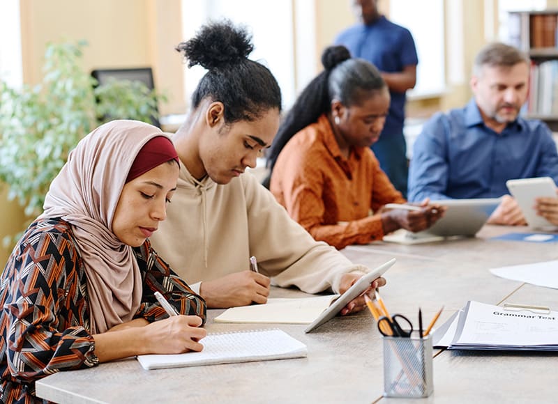 A diverse group of adults is focused on a classroom activity, with some writing notes and others using tablets.