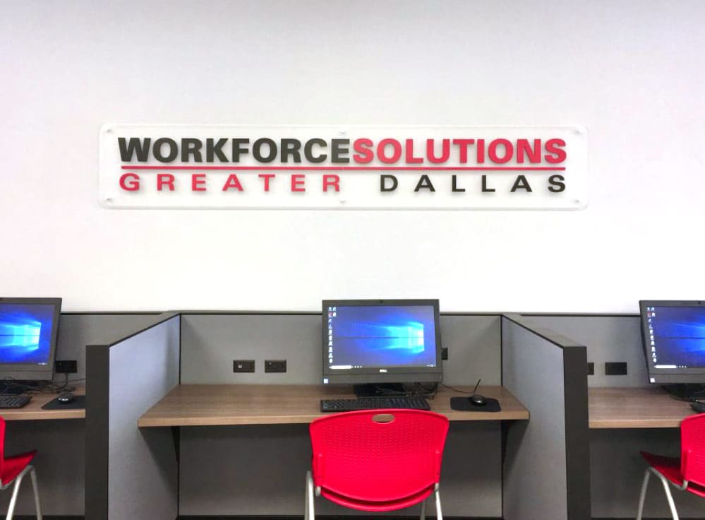 A row of computer workstations with red chairs is set up in a modern office environment. Above the workstations, a sign on the wall reads "Workforce Solutions Greater Dallas" in bold letters. Each workstation has a computer monitor displaying the Windows desktop screen, indicating that the space is designed for public use, likely for job searching or workforce development activities. The setup is clean and organized, reflecting a professional and welcoming atmosphere.